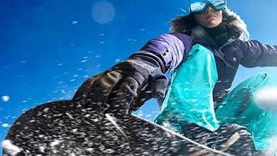 Close-up, low-angle shot of a snowboarder kicking up snow against a bright blue sky on a sunny day.