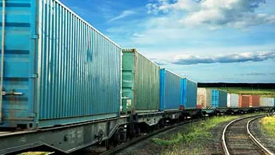 Freight train with blue and green containers rounding a curve through grassy hills under a partly cloudy sky.