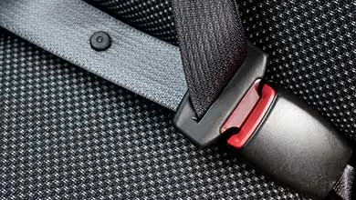 Close-up of a dark gray seatbelt fastened into a black buckle with a red release button on a textured car seat.