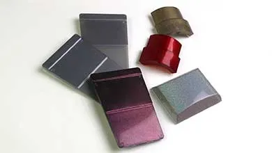 Small molded sample parts in various shapes and metallic finishes—gray, purple, red, and bronze—are scattered on a white surface.