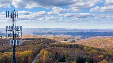 A tall telecommunications tower stands above rolling hills with autumn-colored trees, beneath a blue sky with white clouds.
