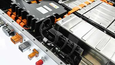 Close-up, angled view of an electric vehicle battery pack, showing silver battery modules in rows, orange high-voltage cables, and a black power distribution unit inside a metal frame.