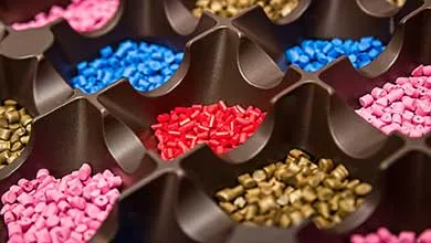 Close-up of a dark brown tray with several compartments, each holding small plastic pellets in pink, red, blue, and gold.