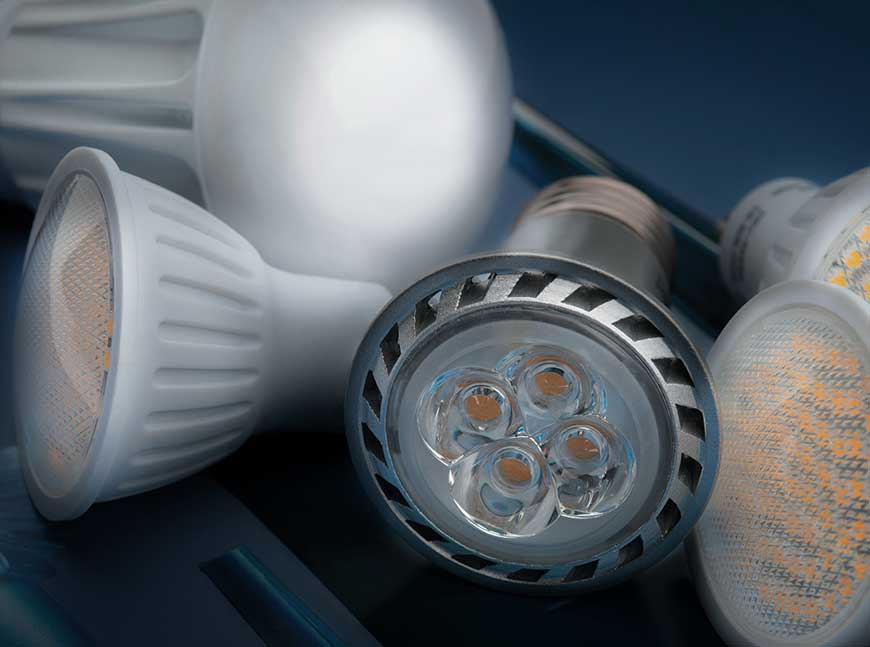 Close-up of assorted LED light bulbs on a dark blue background, highlighting a silver-rimmed spotlight with four visible LED chips in front and white bulbs with clusters of small yellow-orange diodes to the right, with the AVIENT logo in the bottom corner.